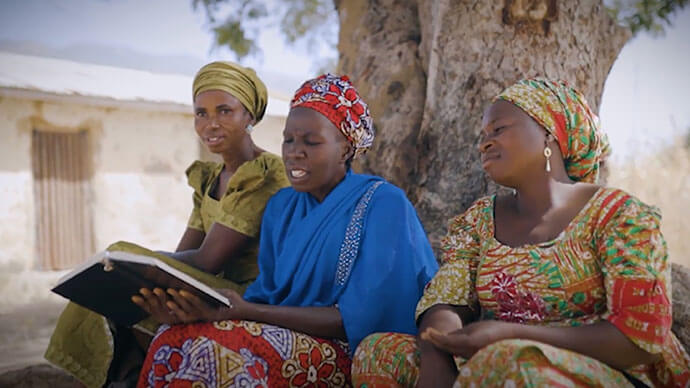 Three African women sitting under a tree while one reads a book